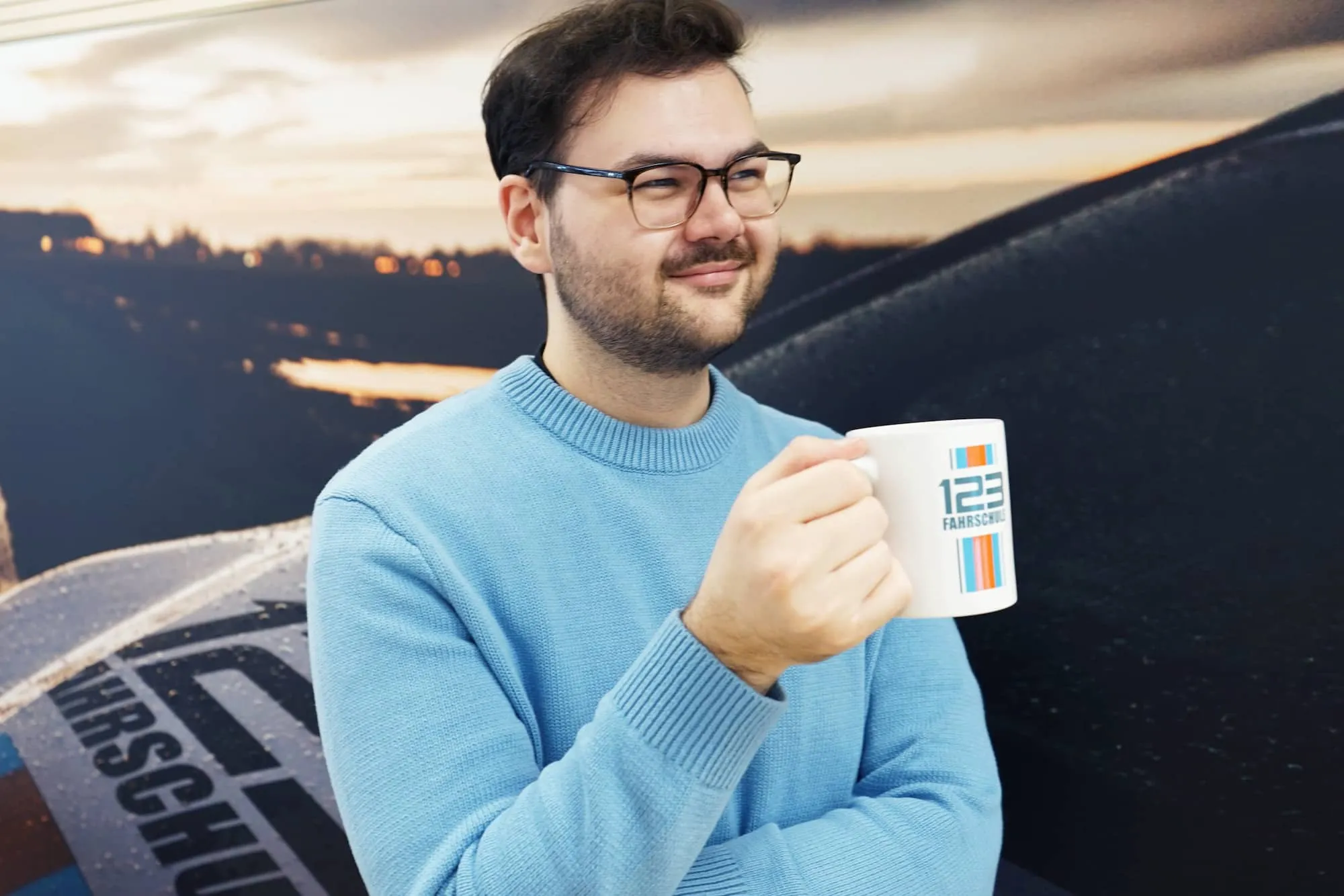 Mann mit Tasse lehnt entspannt an ein Auto einer Fahrschule
